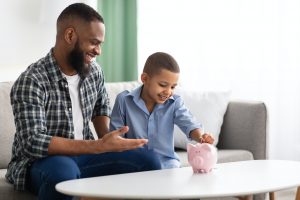 African Father And Son Putting Personal Savings In Piggybank Indoor